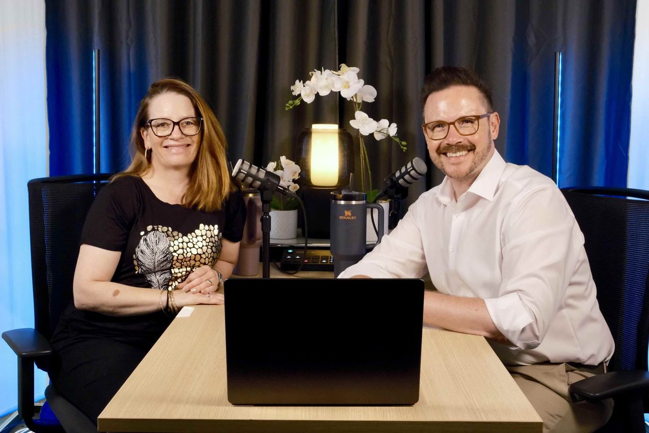 Janine und Oliver sitzen zusammen am Tisch im Podcast-Studio, vor ihnen ein Laptop.